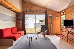 Et værelse med en rød sofa, en træstol, et lille hvidt bord, et fjernsyn på et træskab og en skydedør ud til det fri.
