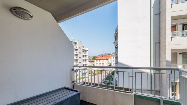 Een balkon met een metalen hekwerk, een kleine vuilnisbak en een uitzicht op gebouwen op de achtergrond.