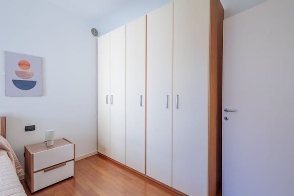 A bedroom with a bed, a nightstand with a lamp, a wardrobe, and a wall-mounted artwork.