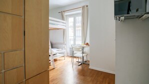 Huoneessa on kerrossänky, ruokapöytä tuoleineen, seinälle asennettu televisio ja ikkuna, jossa on verhot.