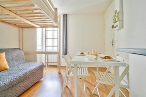 Ein Zimmer mit Holzbalkendecke, einem grauen Sofa, einem weißen Esstisch mit Stühlen und einem Fenster.