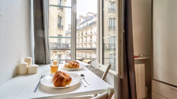 Una mesa de comedor con platos de comida y bebidas, una ventana con vista a edificios y una silla blanca.