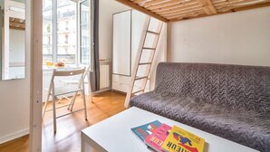 Una habitación con un sofá gris, una mesa blanca con revistas, una silla blanca, una escalera y un ventanal con vistas a edificios.
