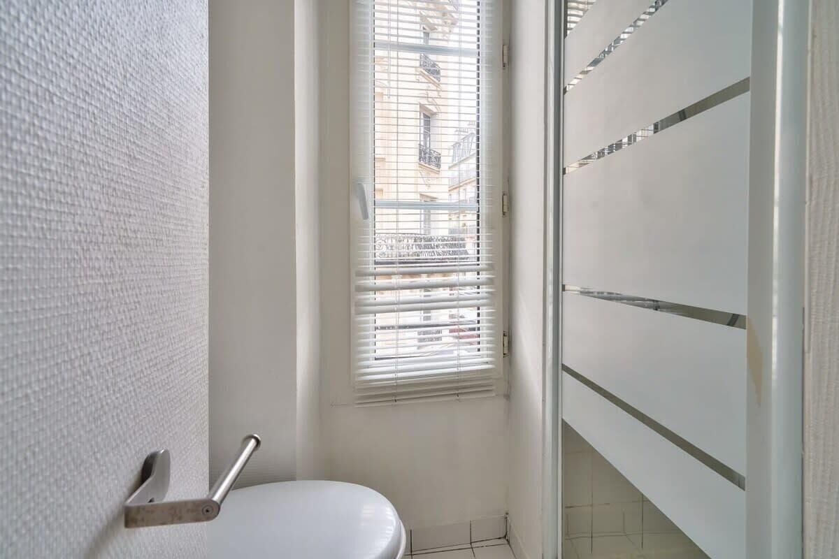 A small, clean bathroom with a toilet, a window with blinds, and a white tiled wall.