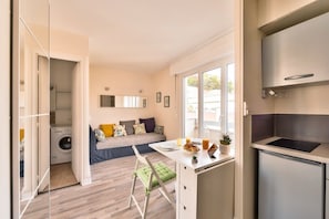A compact apartment with a kitchenette, a dining area, a sofa with cushions, a washing machine, and a window. - Joli Studio, Neuf, Calme & Lumineux -Bail Mobilité (Paris)