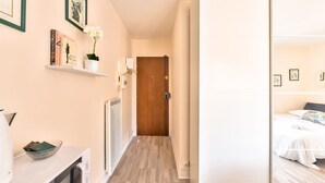 Un pasillo con piso de madera, un radiador blanco, una puerta al fondo y una cama con edredón blanco en la habitación contigua.