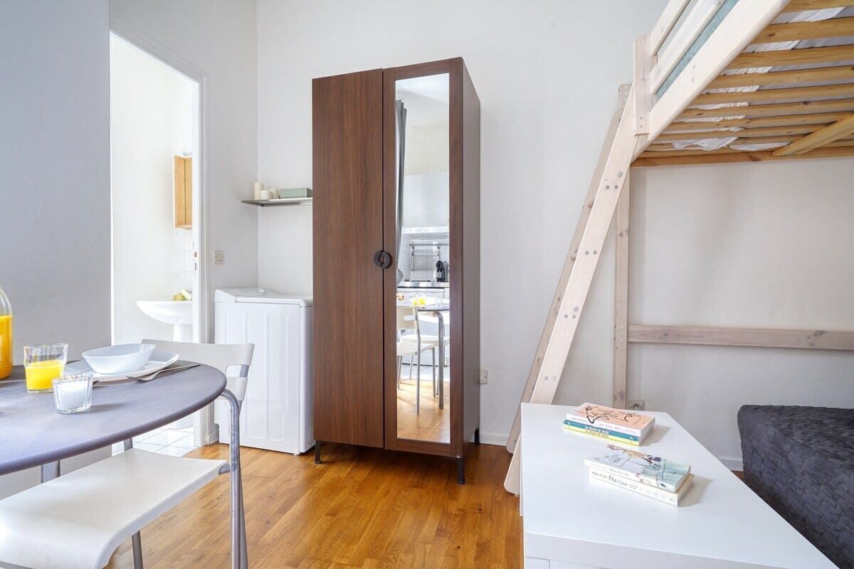 A compact room with a wooden wardrobe, a dining table with chairs, a small kitchenette, and a bed.