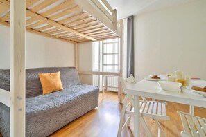 A room with a grey sofa, a white table with breakfast items, and a window with natural light. - Joli studio-Coeur du 17e-Bail Moyenne Durée (1DD) (Paris)