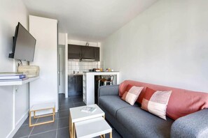 Ett modernt vardagsrum med en grå soffa, en platt-TV som är monterad på väggen, ett litet vitt bord och en köksdel i bakgrunden.