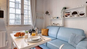 Una habitación con un sofá azul, una mesa blanca con productos para el desayuno, una ventana con cortinas y una estantería con adornos.
