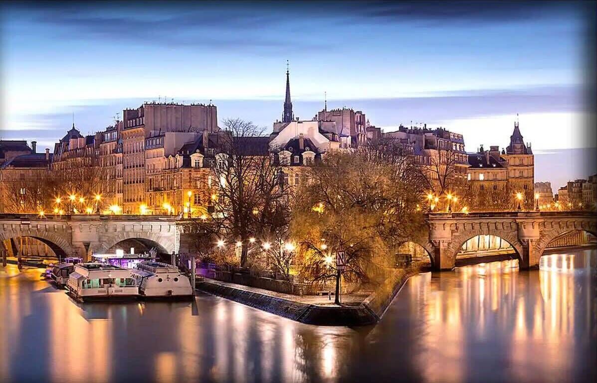 A river with a boat docked on the left, a bridge in the center, and buildings in the background.