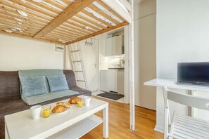 A modern living room with a sofa, a coffee table with breakfast items, a TV on a stand, and a kitchen area visible through an open door. - Studio Parfait - Coeur du 17em (Paris)
