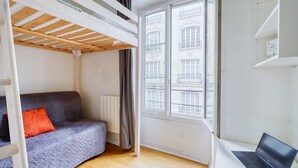 A room with a bunk bed, a sofa with an orange pillow, a laptop on a table, and a window with a view of buildings. - Welcoming Studio - Coeur du 17em - Bail moy durée (Paris)
