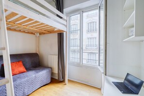 Ein Zimmer mit einem Etagenbett, einem Sofa mit orangefarbenem Kissen, einem Laptop auf einem Tisch und einem Fenster mit Blick auf Gebäude.