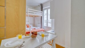 A room with a bunk bed, a dining table with breakfast items, and a window with curtains. - Welcoming Studio - Coeur du 17em - Bail moy durée (Paris)