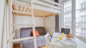 A room with a bunk bed, a gray sofa, a dining table with breakfast items, and a window with a view of buildings. - Welcoming Studio - Coeur du 17em - Bail moy durée (Paris)