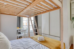 A bedroom with a wooden ceiling, a bed with a yellow blanket, a white chair, a desk with a chair, and a window. - Nicely designed Studio - Paris 17 - Mid Terms (Paris)