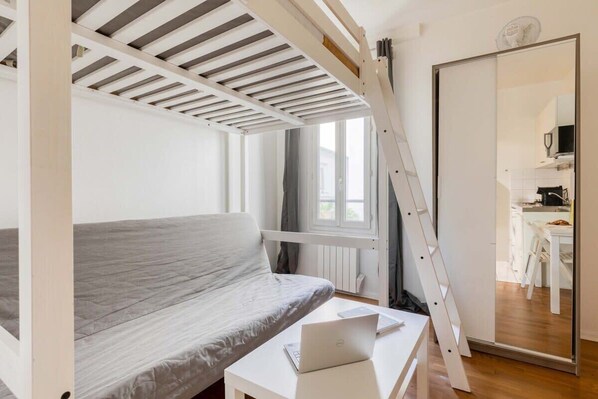 Ein Schlafzimmer mit einem Bett, einem Schreibtisch mit Laptop, einer Leiter und Blick auf eine Küche durch eine Schiebetür.