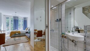 A modern hotel room with a large bed, a sitting area with a sofa and chairs, a dining table, a shower area with a glass door, and a bathroom with a sink and mirror. - New, Large and Cosy Studio - Le Marais - Mid Terms (Paris)