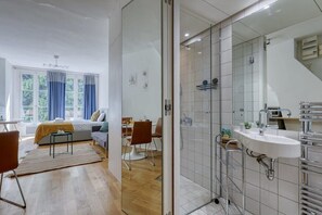 A modern hotel room with a large bed, a sitting area with a sofa and chairs, a dining table, a shower area with a glass door, and a bathroom with a sink and mirror. - New, Large and Cosy Studio - Le Marais - Mid Terms (Paris)