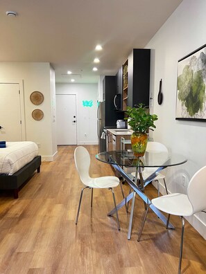 Ein moderner Wohnraum mit Esstisch aus Glas, weißen Stühlen, einem Bett, einer Küchenzeile und einem Wandbild.