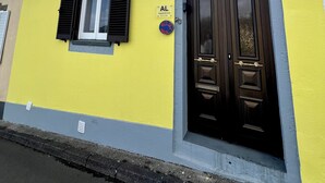 Um edifício amarelo com porta preta e portaria, um letreiro com a inscrição 'AL', e uma parede azul e branca.