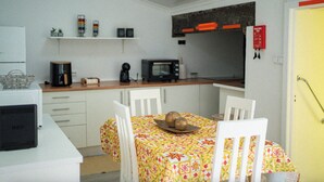 Cozinha compacta com armários brancos, micro-ondas e cafeteira. Há uma mesa de jantar com toalha de mesa estampada e cadeiras brancas.