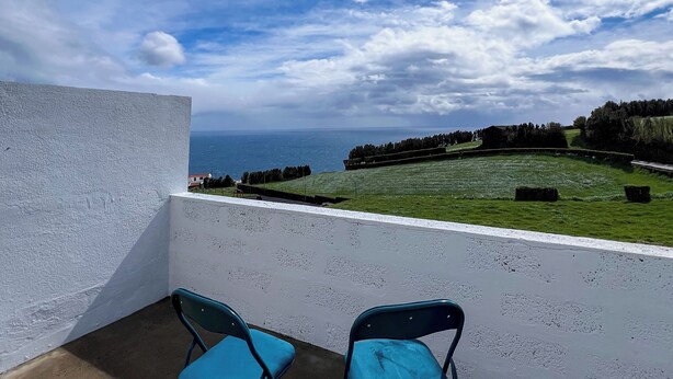 Uma varanda com duas cadeiras azuis, uma parede branca e vista para o mar e a paisagem.
