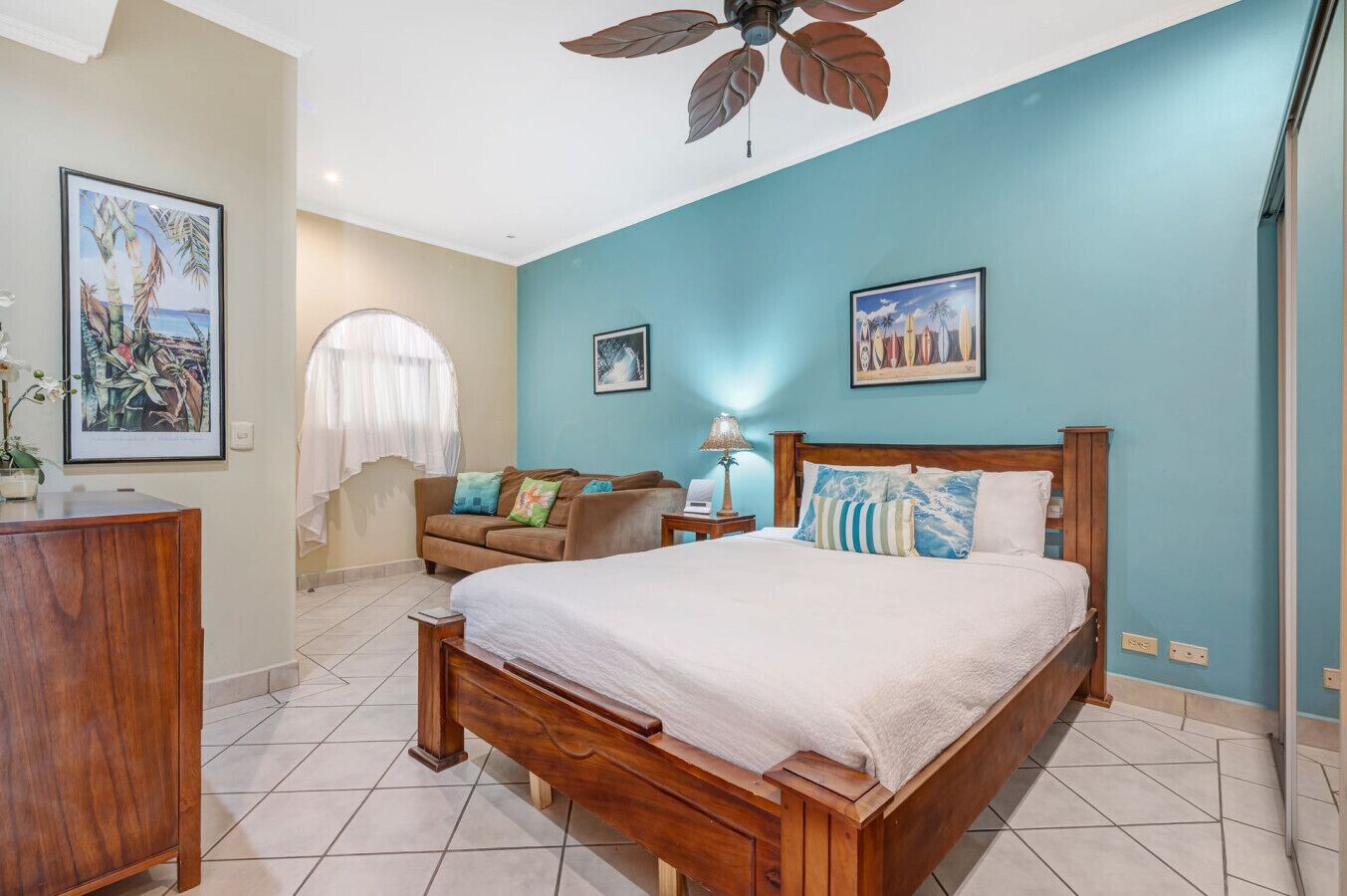 A bedroom with a wooden bed, a nightstand, a lamp, a window with white curtains, and a ceiling fan.