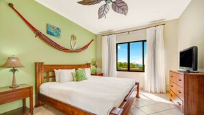 A bedroom with a bed, a wooden headboard, a nightstand with a lamp, a dresser, a television, a hammock, a ceiling fan, and a window with curtains. - Sunrise Tamarindo 22 (Tamarindo)