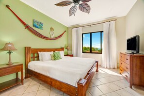 Ein Schlafzimmer mit Bett, Holz-Kopfstück, Nachttisch mit Lampe, Kommode, Fernseher, Hängematte, Deckenventilator und Fenster mit Vorhängen.