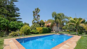 Une piscine avec une terrasse carrelée, entourée de verdure et une maison en arrière-plan.