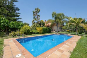 En swimmingpool med fliseterrasse, omgivet af grønt og et hus i baggrunden.