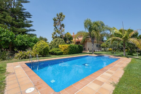Une piscine avec une terrasse carrelée, entourée de verdure et une maison en arrière-plan.