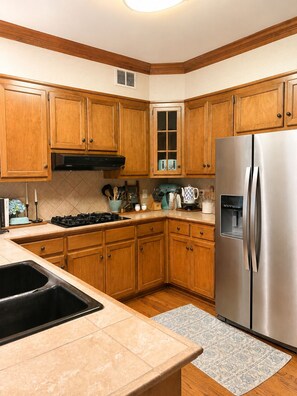 A kitchen with wooden cabinets, a stainless steel refrigerator, a gas stove, and a window with blinds. - Relaxing Masters Week Getaway Near Savannah River & Evans 20 Min to the National (Evans)