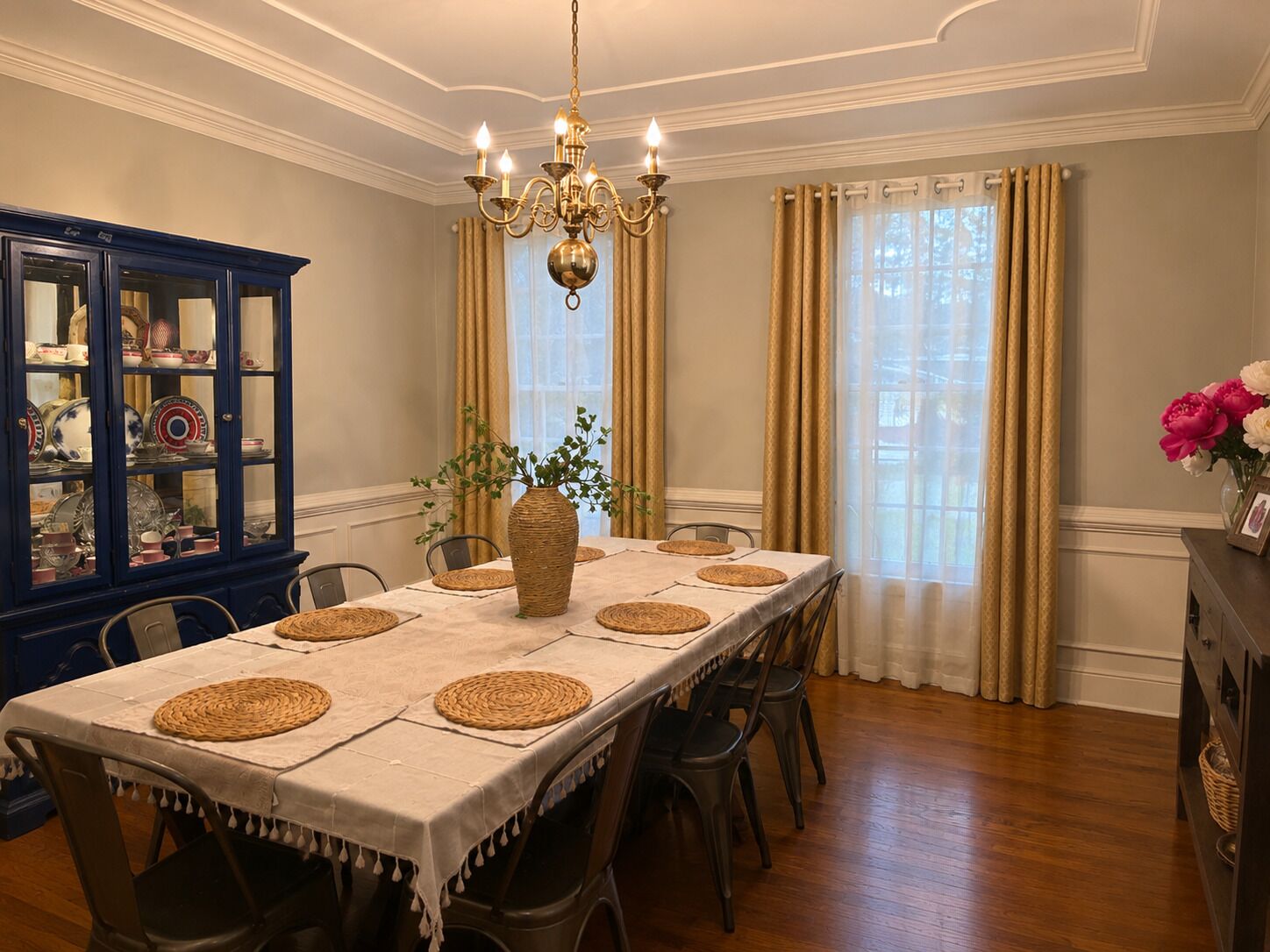 A dining room with a long table, chairs, a chandelier, a cabinet with dishes, and curtains.