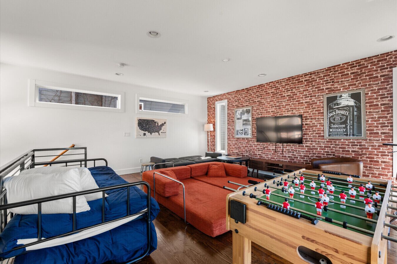 A room with bunk beds, a sofa, a foosball table, a brick wall, and a mounted TV.