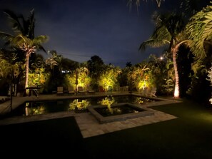 Un área de piscina nocturna con sillones de descanso y palmeras.