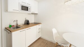 Une cuisine moderne avec des meubles blancs, un évier en acier inoxydable, un four à micro-ondes et une machine à café. On y trouve une chaise blanche et une table ronde.