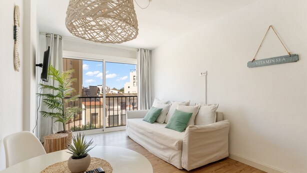 Un salon avec un canapé blanc, une table ronde sur laquelle se trouve une télécommande, une plante en pot et un balcon avec vue.