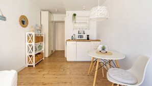 Une cuisine compacte avec des meubles blancs, un coin repas doté d’une table et de chaises blanches, et un luminaire suspendu.