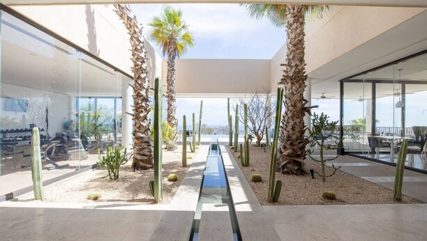 A modern indoor space with a central water feature, surrounded by cacti and palm trees, with large windows offering views. - Condo with Arco views, infinity pool, and spa (Cabo San Lucas)