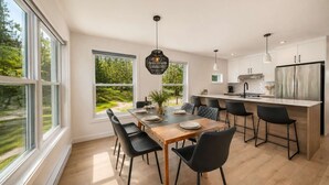 A modern kitchen with large windows, a dining area with a wooden table and black chairs, and a stainless steel refrigerator. - Spa & Nature Escape | Le Nova (Adstock)