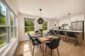 A modern kitchen with large windows, a dining area with a wooden table and black chairs, and a stainless steel refrigerator.