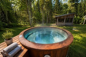 A hot tub in a forested area with a wooden deck, a small cabin, and a potted plant.