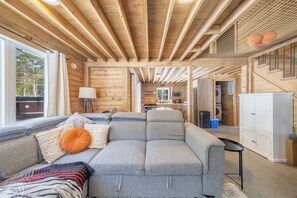 A log cabin interior with a sofa, a lamp, a kitchen area, and a staircase.