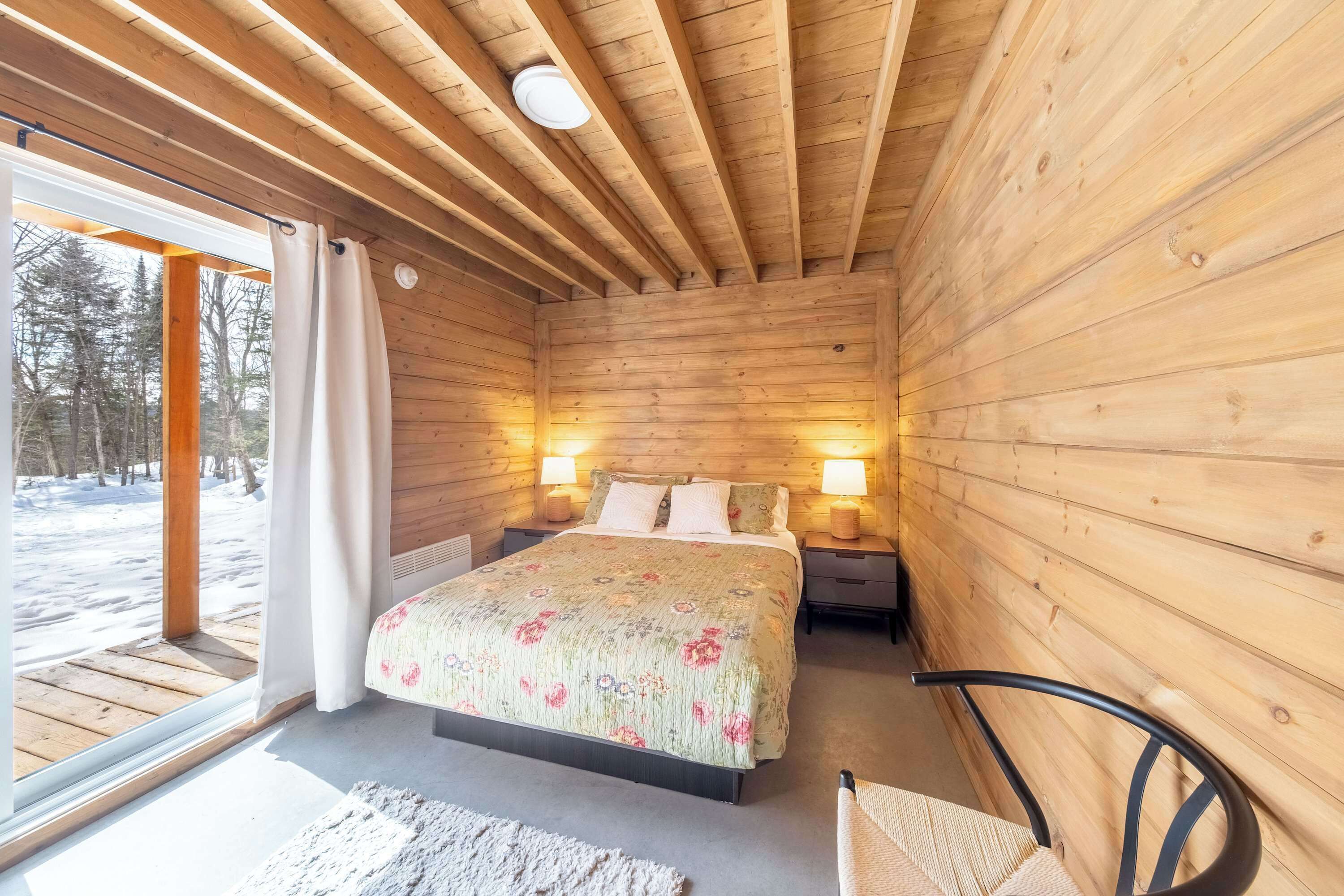 Un dormitorio de cabaña de madera con una cama, lámparas de noche y un ventanal con cortinas.