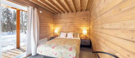 Un dormitorio de cabaña de madera con una cama, lámparas de noche y un ventanal con cortinas.