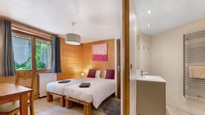 Slaapkamer met twee bedden, een houten vloer, een raam met gordijnen, een nachtkastje, een lamp en een badkamer met een wastafel en handdoekrek.