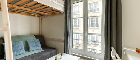 Una habitación con literas, un sofá, una mesita con plantas y una ventana con vistas a edificios.